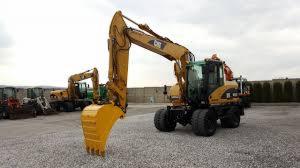 Żółta koparka kołowa Caterpillar z łyżką opuszczoną do ziemi na żwirowym placu, w tle druga koparka i betonowy mur.