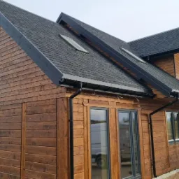 Nowoczesny, dwukondygnacyjny dom z ciemnego drewna z czarną dachówką bitumiczną i oknami w grafitowych ramach, widok z zewnątrz na elewację z widoczną fakturą drewna i fragmentem tarasu.