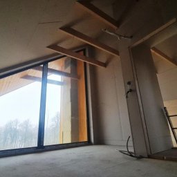 RIWKE - Wnętrze domu szkieletowego w trakcie budowy, widoczne drewniane elementy konstrukcyjne, okno z widokiem na las oraz drabina oparta o ścianę.