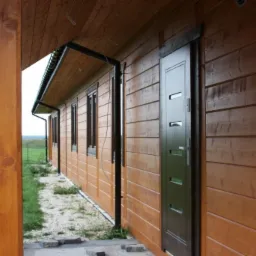 Zewnętrzny widok brązowego domu z drewnianą elewacją, ciemnymi oknami i drzwiami, oraz czarnymi rynnami.