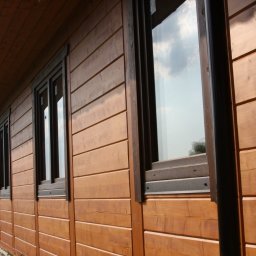 RIWKE - Zewnętrzna ściana domu szkieletowego z drewnianą elewacją, widoczne okna z ciemnymi ramami i rynna, z perspektywy bocznej.