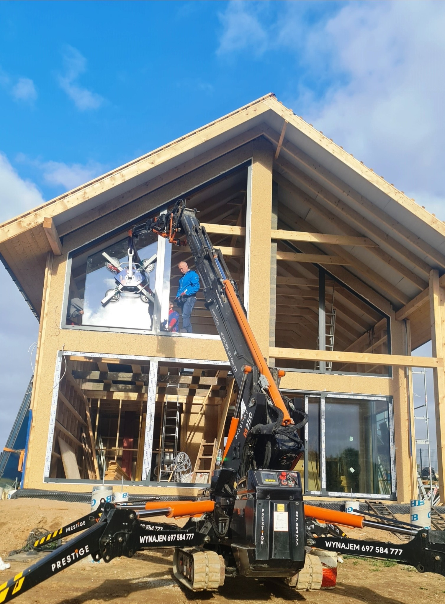 Montaż okna w surowym stanie domu szkieletowego z użyciem dźwigu z przyssawkami, widoczna drewniana konstrukcja i pracownik w niebieskiej kurtce.