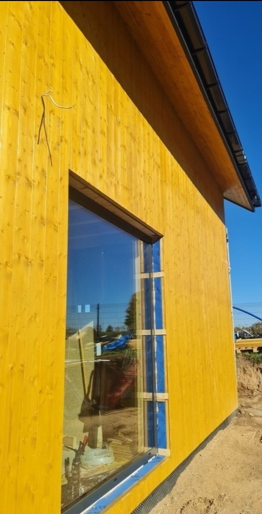 Żółta elewacja domu szkieletowego z dużym oknem, zabezpieczonym niebieską folią, widoczne narzędzia wewnątrz budynku, niepodłączone kable na zewnątrz.