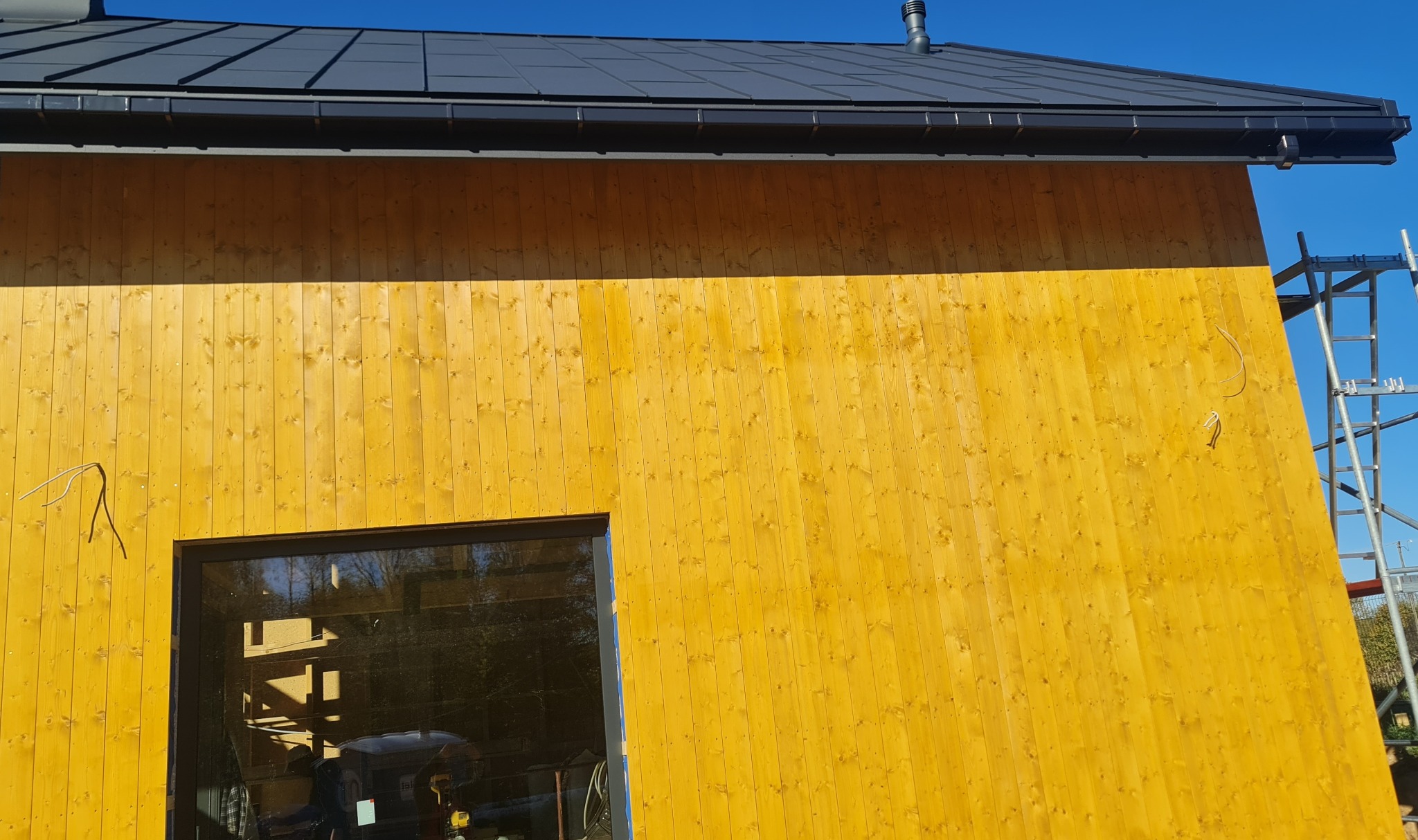 Fragment ściany zewnętrznej budynku z jasnego drewna z widocznym oknem i elementami instalacji elektrycznej oraz rusztowaniem w tle.