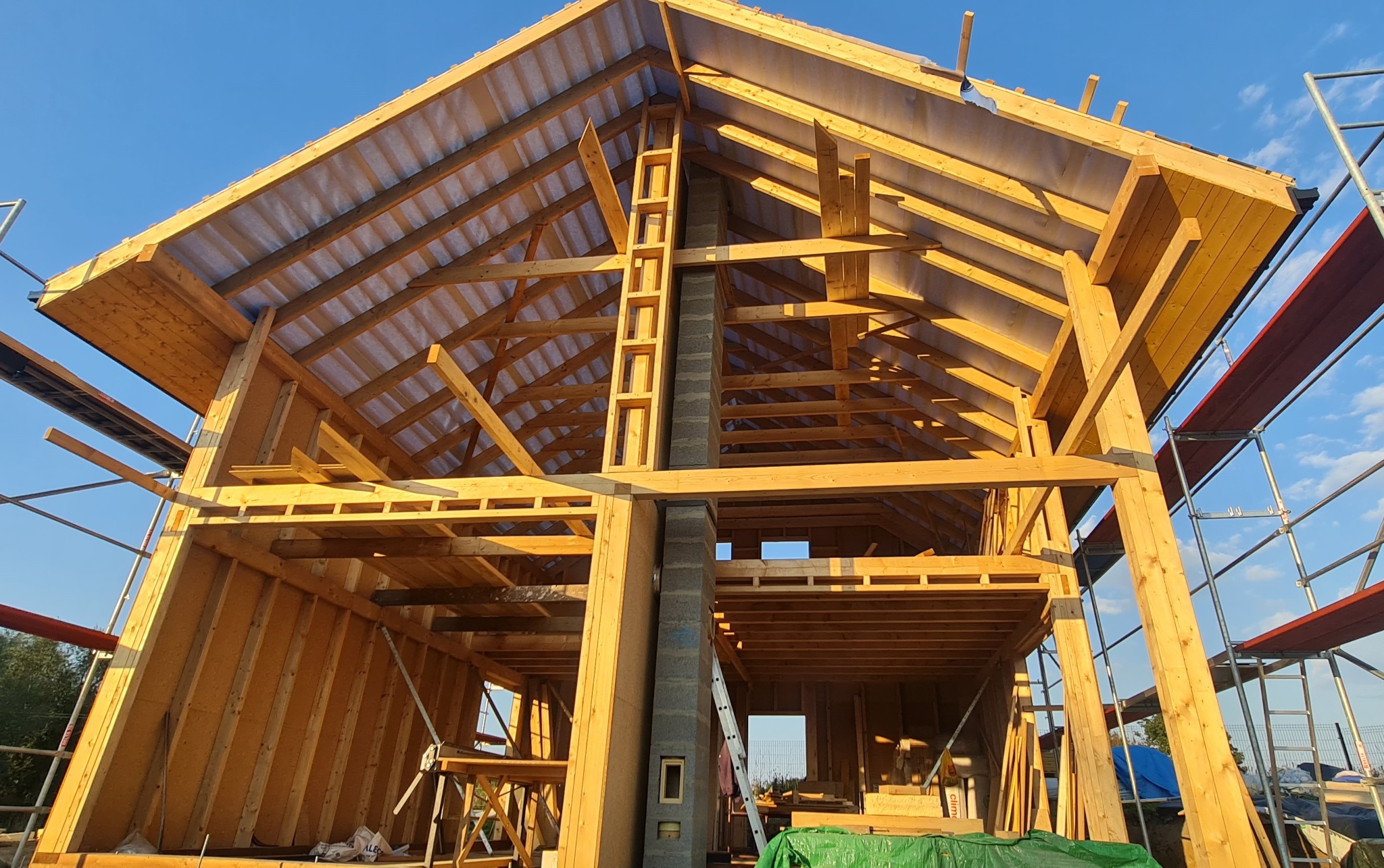 Surowa konstrukcja domu z drewnianych belek i szarego komina z pustaków, widok z dołu na tle błękitnego nieba, na rusztowaniach czerwone deski.