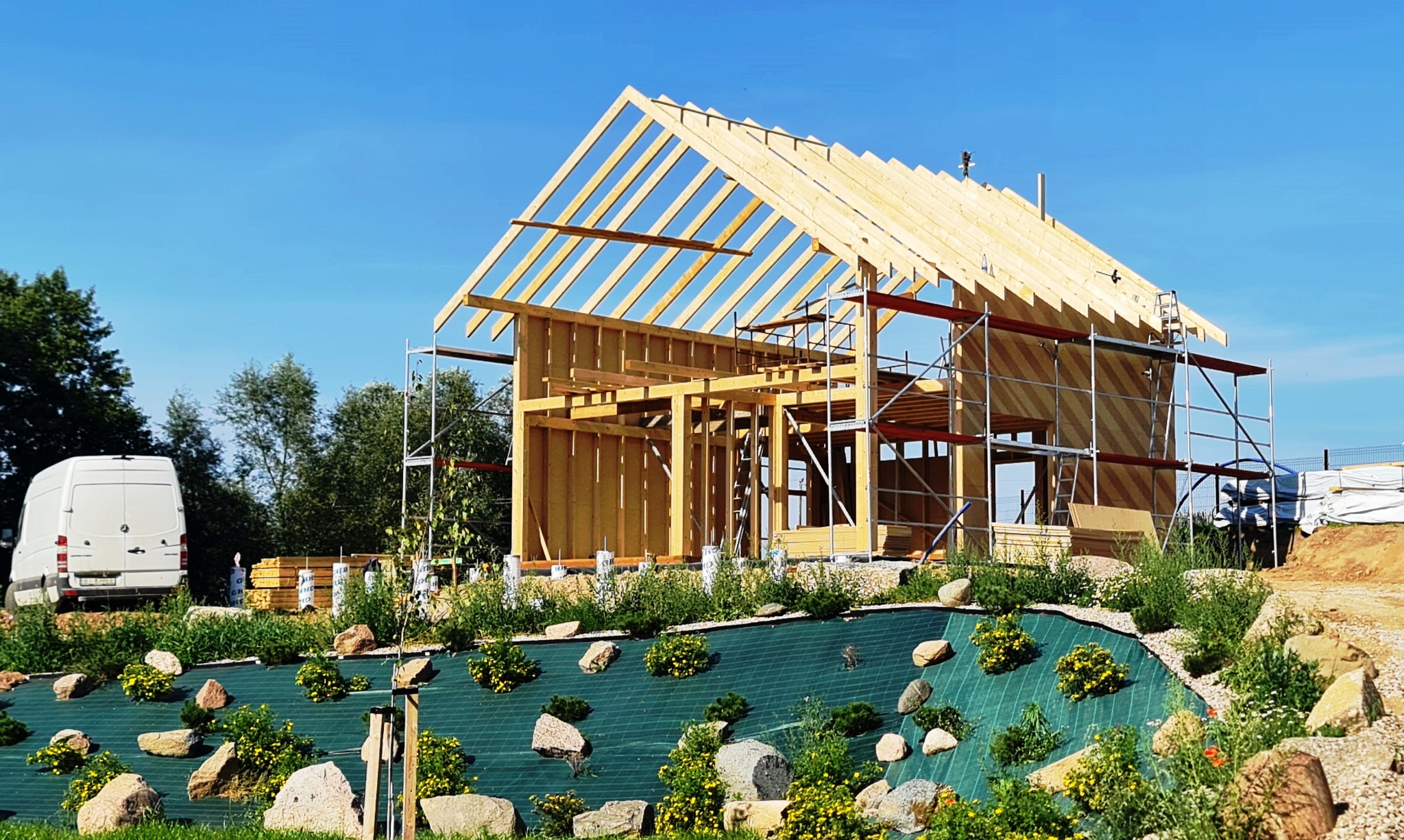 Wznoszenie konstrukcji domu z drewnianych belek na fundamencie, z widocznymi rusztowaniami i dostawczym busem w tle, otoczone zielenią i kamieniami.
