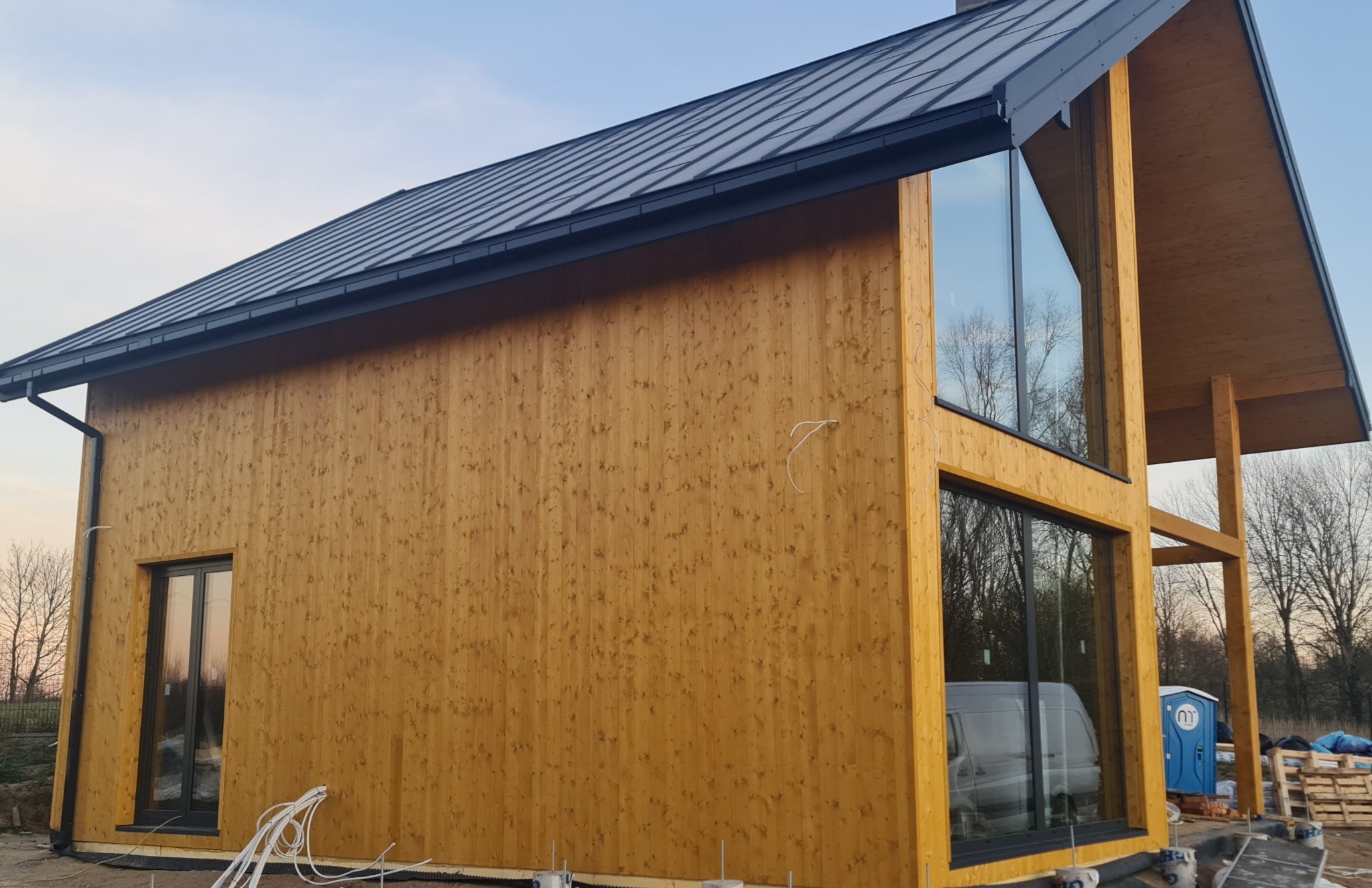 Nowoczesny dom z drewnianą elewacją i dużymi oknami, widoczne kable na zewnątrz, w tle las i niebieska toaleta przenośna.