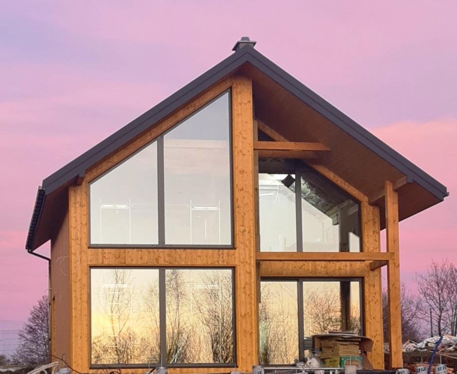 Surowy drewniany dom z dużymi oknami na tle różowego nieba o zachodzie słońca, widoczne rusztowania i materiały budowlane u podstawy.