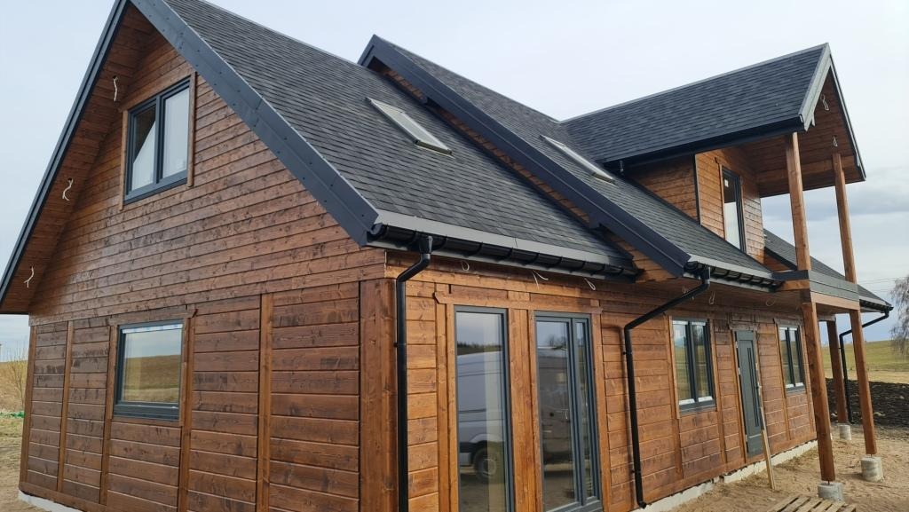 Nowoczesny, dwukondygnacyjny dom z ciemnego drewna z czarną dachówką bitumiczną i oknami w grafitowych ramach, widok z zewnątrz na elewację z widoczną fakturą drewna i fragmentem tarasu.