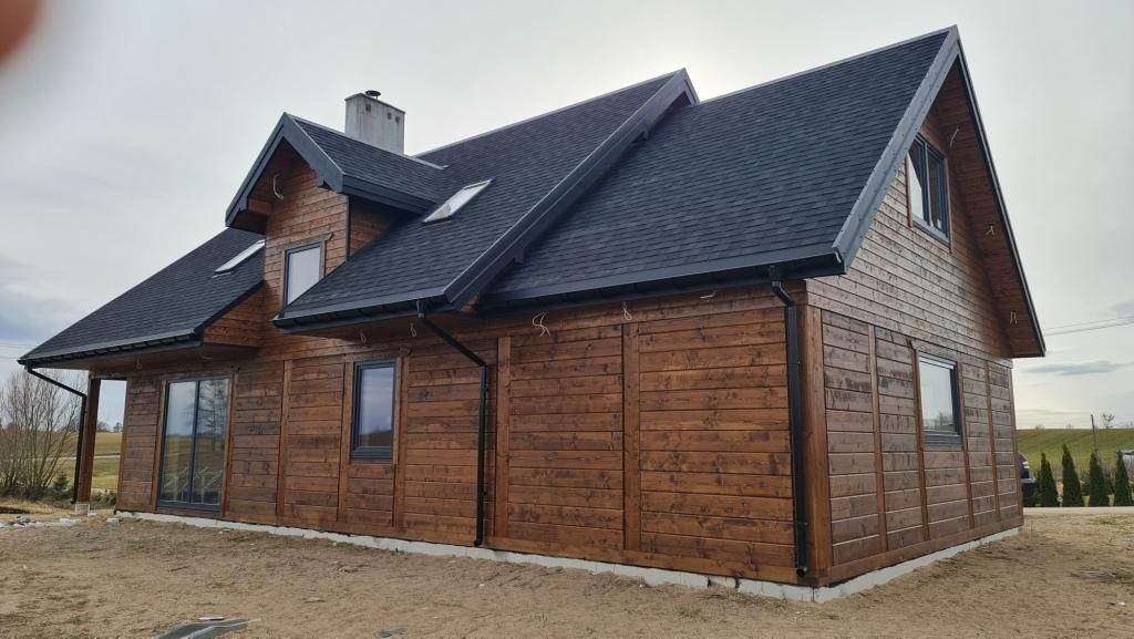 Gotowy, parterowy dom z brązowych, impregnowanych desek z ciemnym dachem i kominem, widok z boku na tle pochmurnego nieba.