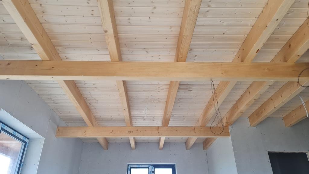 Konstrukcja drewnianego stropu z belek i desek podbiciowych w surowym wnętrzu, widoczne okno i wystające przewody elektryczne.