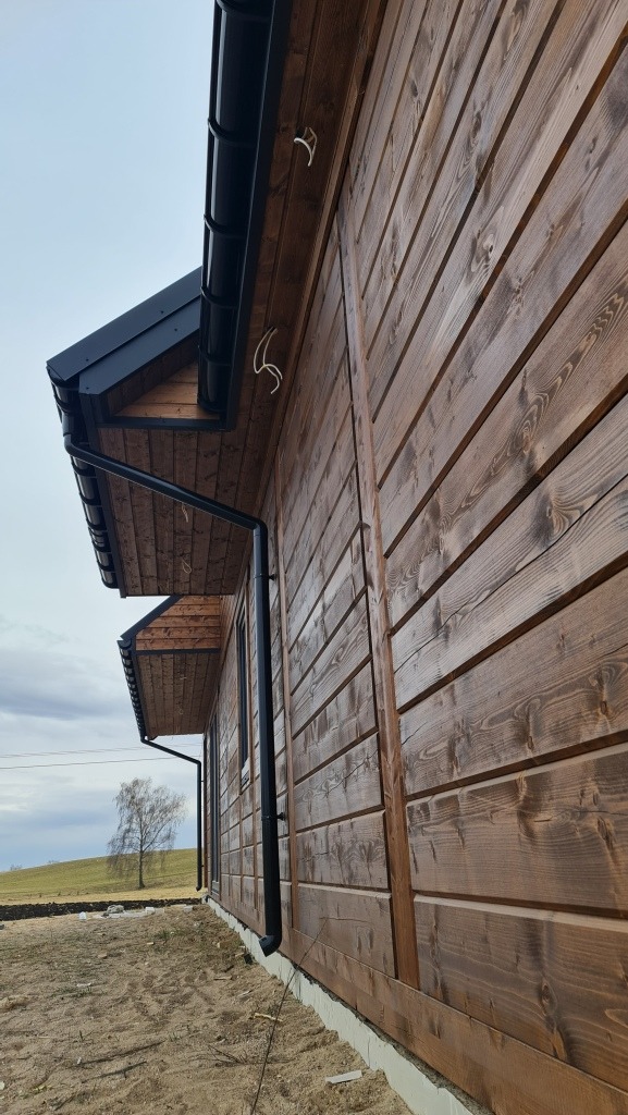 Fragment elewacji domu z ciemnego drewna, widoczne czarne rynny i uchwyty, w tle zielone wzgórze i samotne drzewo.
