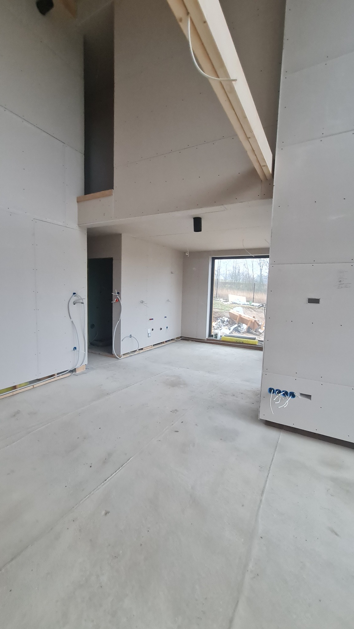 Wnętrze domu w trakcie budowy: surowe ściany z płyt gipsowo-kartonowych, widoczne instalacje elektryczne i sanitarne, betonowa podłoga, drewniane belki stropowe, okno z widokiem na plac budowy.