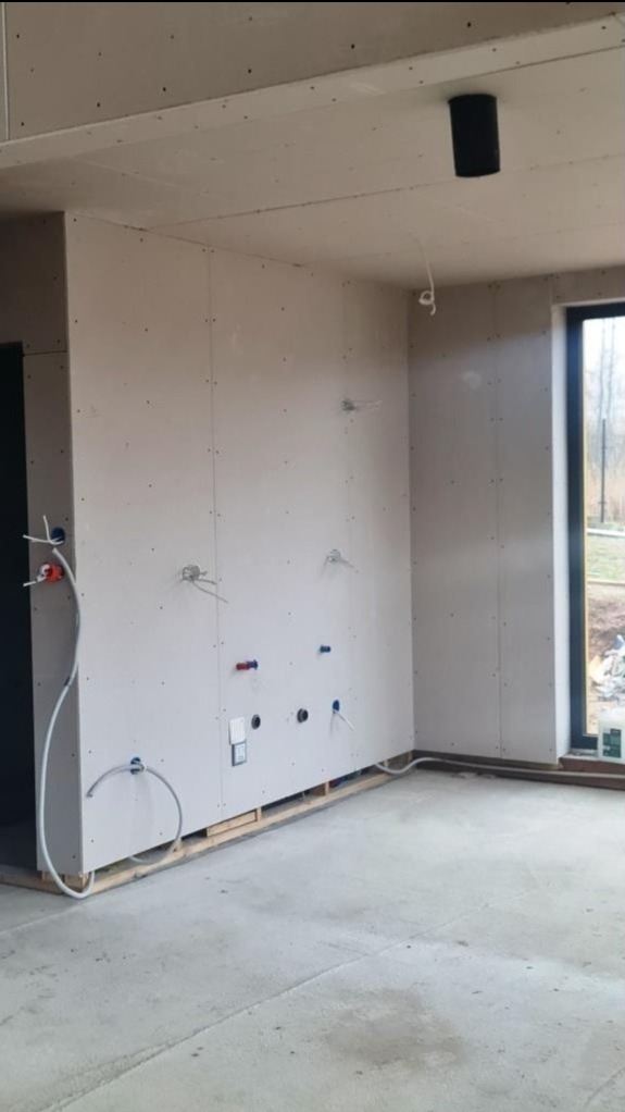 Wnętrze w trakcie wykańczania: ściany z płyt gipsowo-kartonowych, widoczne instalacje elektryczne i hydrauliczne, podłoga betonowa, czarna tuba wentylacyjna pod sufitem, okno z widokiem na zewnątrz.