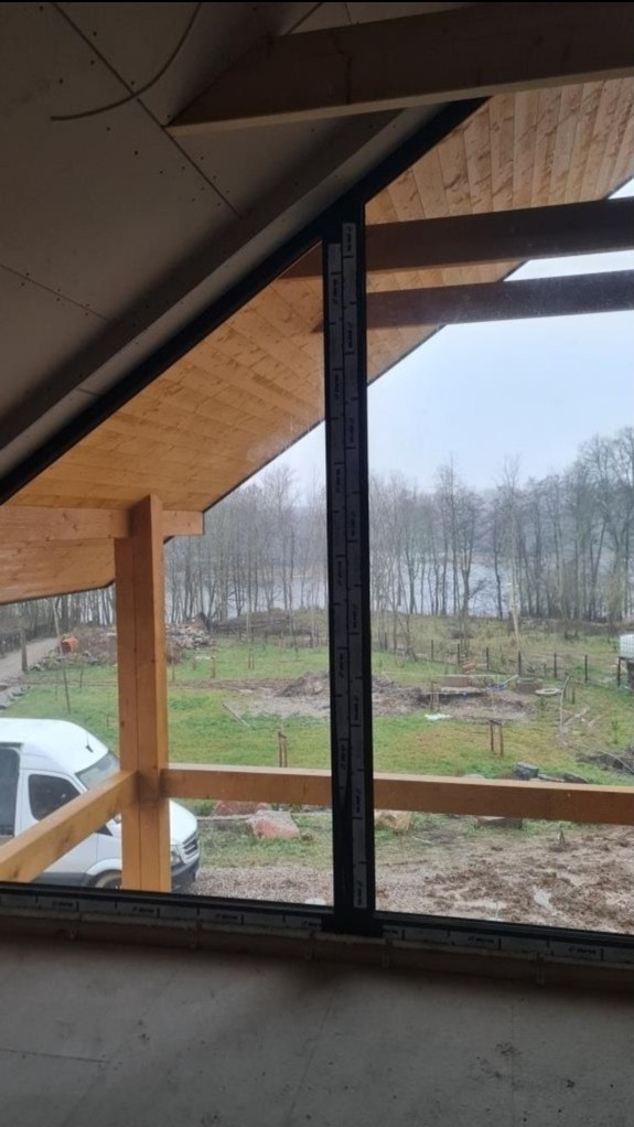Wnętrze domu w trakcie budowy z drewnianą konstrukcją dachu i dużym oknem z widokiem na krajobraz z jeziorem w tle, widoczny biały samochód dostawczy na zewnątrz.