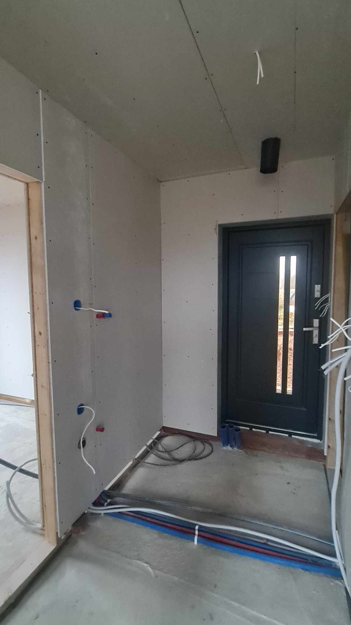 Wnętrze w trakcie wykańczania: ściany z płyt gipsowo-kartonowych, instalacja elektryczna i hydrauliczna w podłodze, czarne drzwi wejściowe z przeszkleniami.