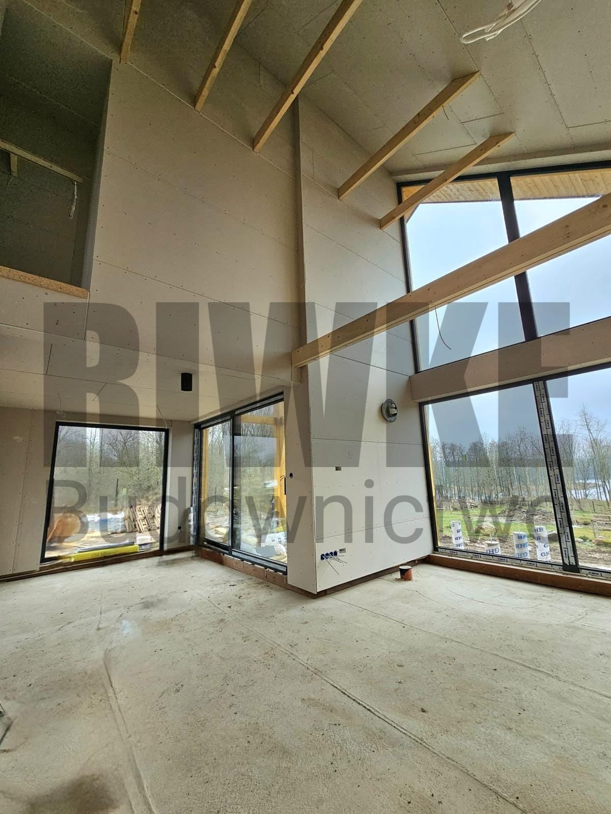 Wnętrze domu w trakcie budowy, widoczne surowe ściany z płyt, drewniane belki stropowe, duże okna z widokiem na drzewa i jezioro, instalacja elektryczna w ścianie.