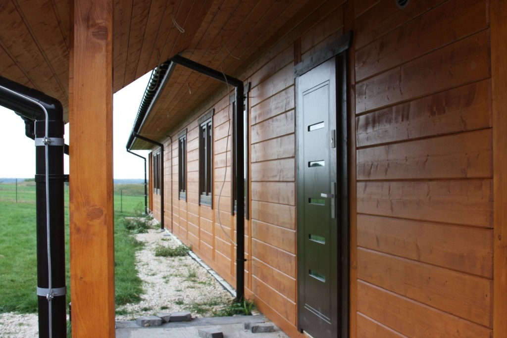 Zewnętrzny widok brązowego domu z drewnianą elewacją, ciemnymi oknami i drzwiami, oraz czarnymi rynnami.