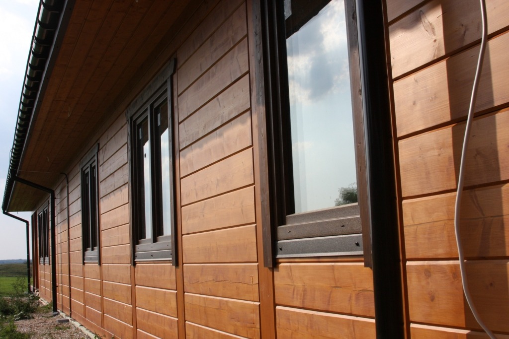 Zewnętrzna ściana domu szkieletowego z drewnianą elewacją, widoczne okna z ciemnymi ramami i rynna, z perspektywy bocznej.