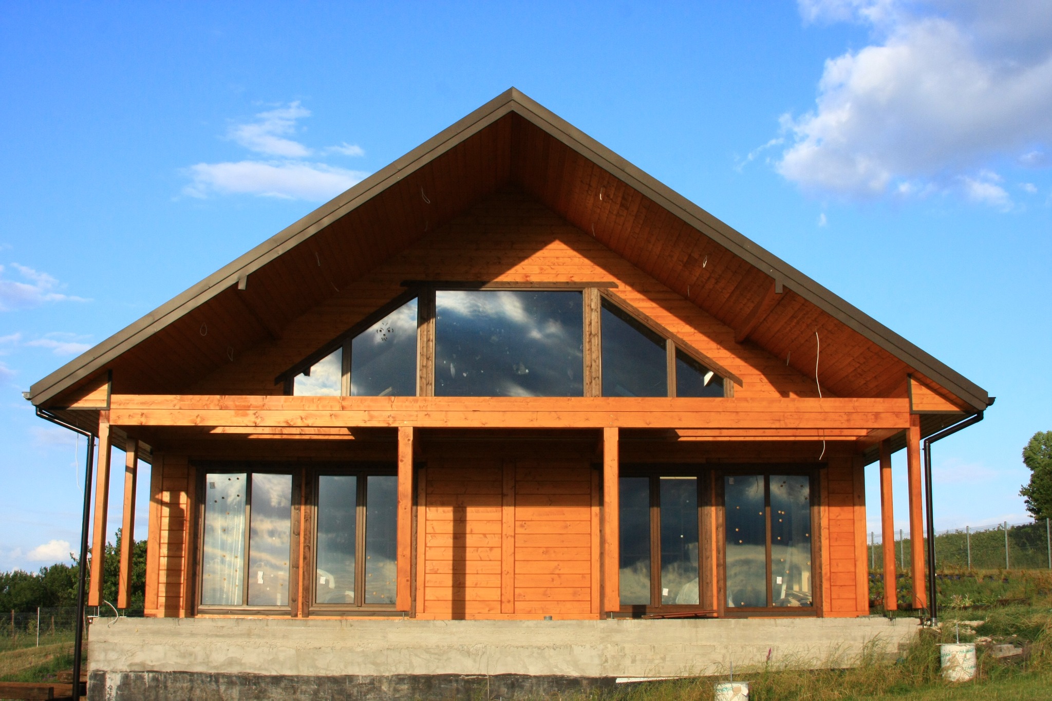 Surowy stan drewnianego domu z dużymi oknami i trójkątnym świetlikiem, na fundamencie z betonu, z widocznym niebieskim niebem z chmurami.