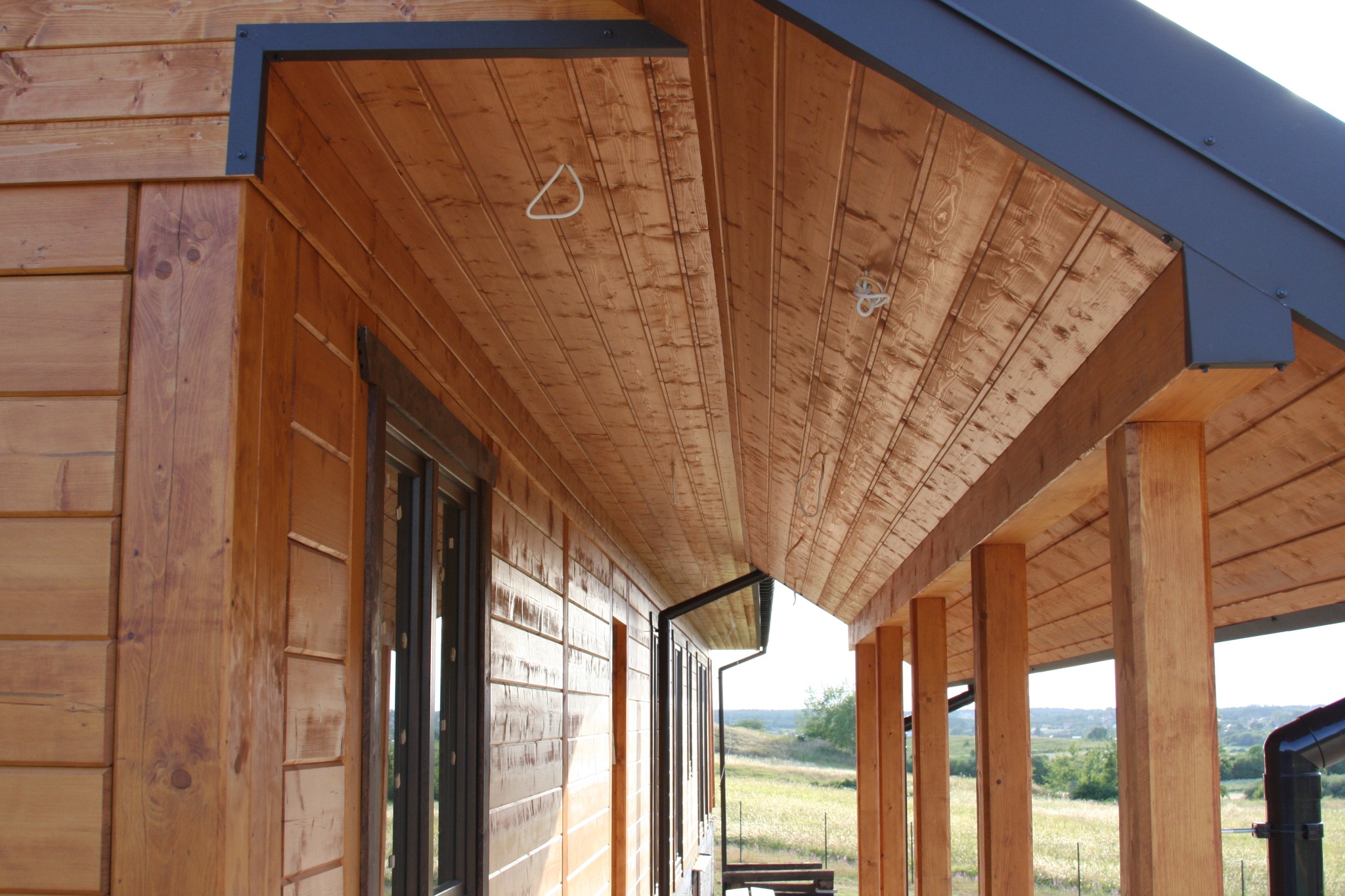 Drewniana elewacja domu z widocznym zadaszeniem tarasu, bejcowane deski, fragment okna i słupy podtrzymujące dach.