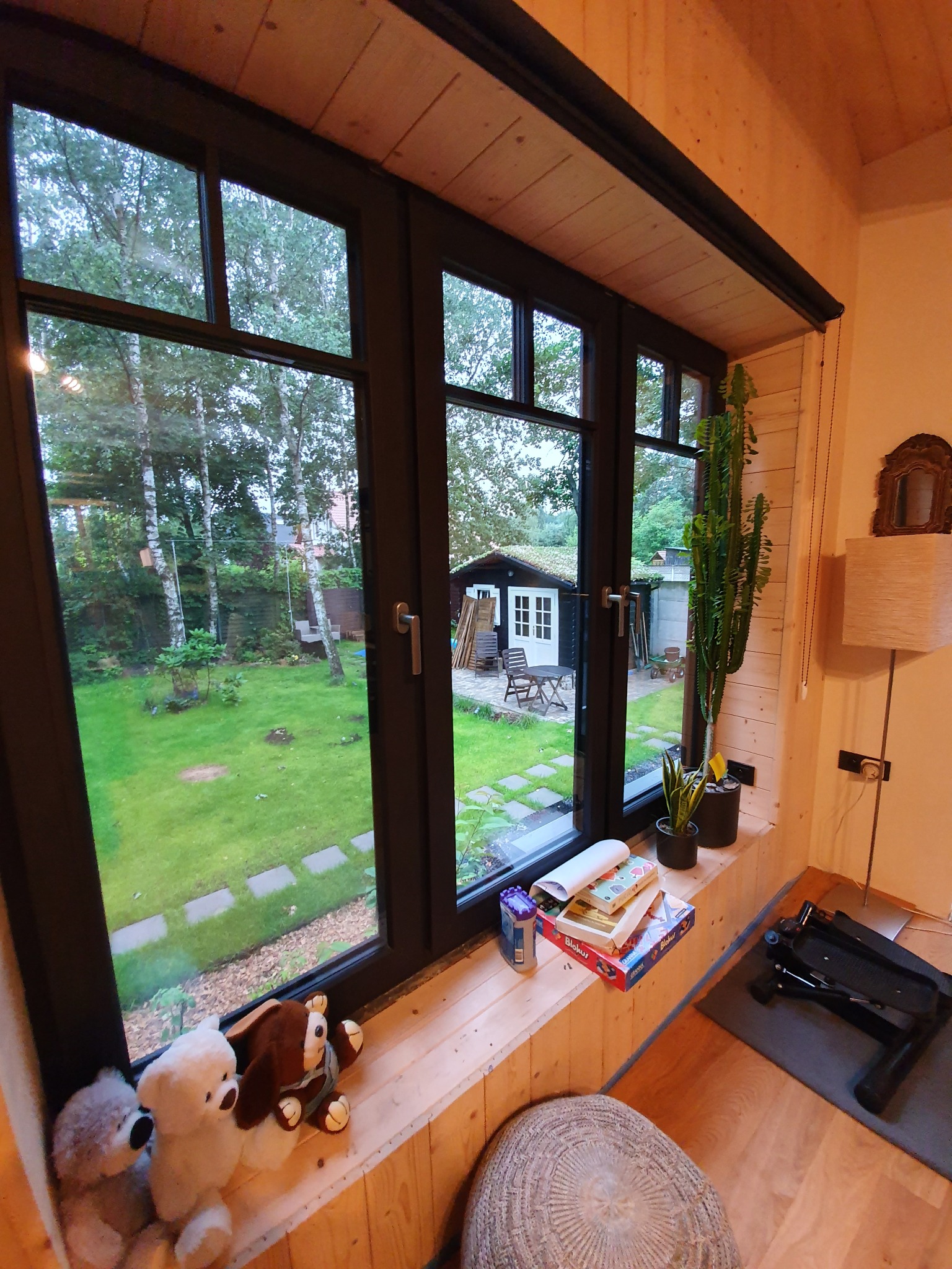 Wnętrze drewnianego domu z dużym oknem z widokiem na ogród z domkiem narzędziowym, na parapecie pluszowe zabawki, gry planszowe i doniczki z roślinami.