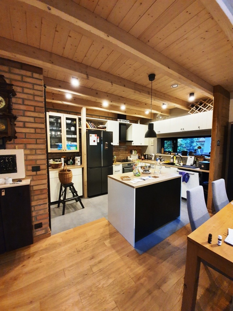 Jasna, przestronna kuchnia w domu z drewnianym stropem belkowym, z wyspą kuchenną w centrum i ceglanym kominem w rogu, widok z perspektywy podłogi z desek.