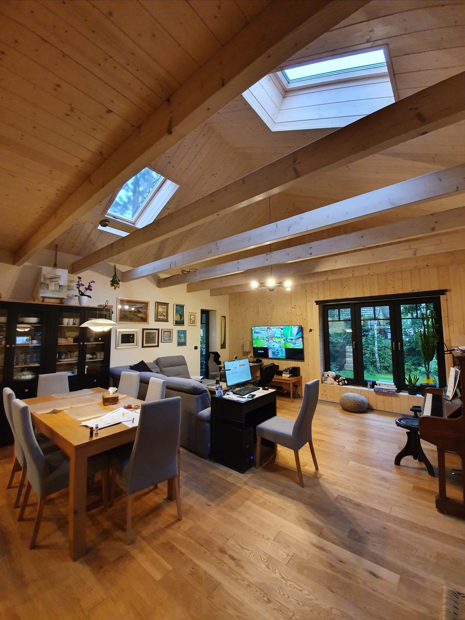 Jasne wnętrze domu w Białymstoku z drewnianym stropem, belkami i oknami dachowymi. Widoczny stół z krzesłami, kanapa, szafka, pianino oraz okno z widokiem na ogród.