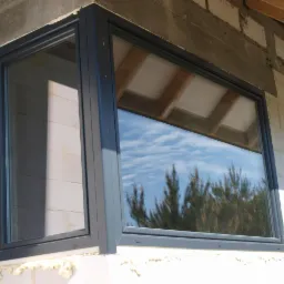 Nowe okno z szarą ramą, zamontowane w ścianie z bloczków, z widocznym odbiciem drzew i nieba w szybie.