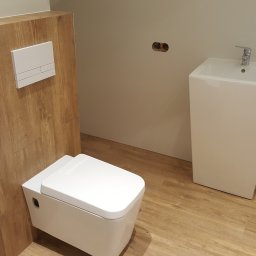 QUEST Firma remontowo budowlana - Nowoczesna, podwieszana toaleta z drewnianą obudową i minimalistyczną umywalką w łazience wykończonej panelami podłogowymi imitującymi drewno.
