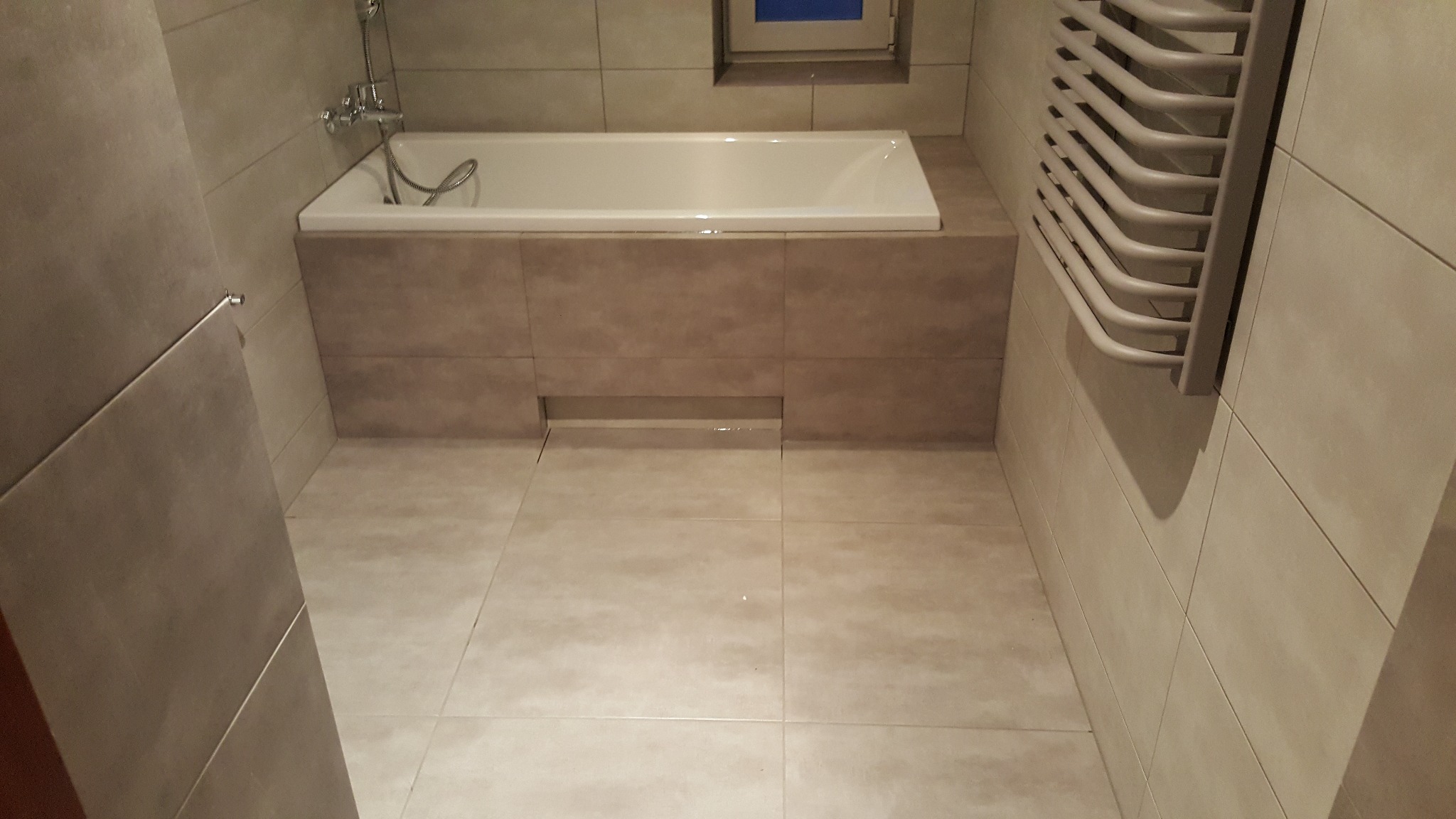 Beżowa łazienka z wanną obudowaną płytkami, grzejnikiem drabinkowym i podłogą wyłożoną kafelkami. Minimalistyczny design, stonowana kolorystyka, widoczny odpływ liniowy.