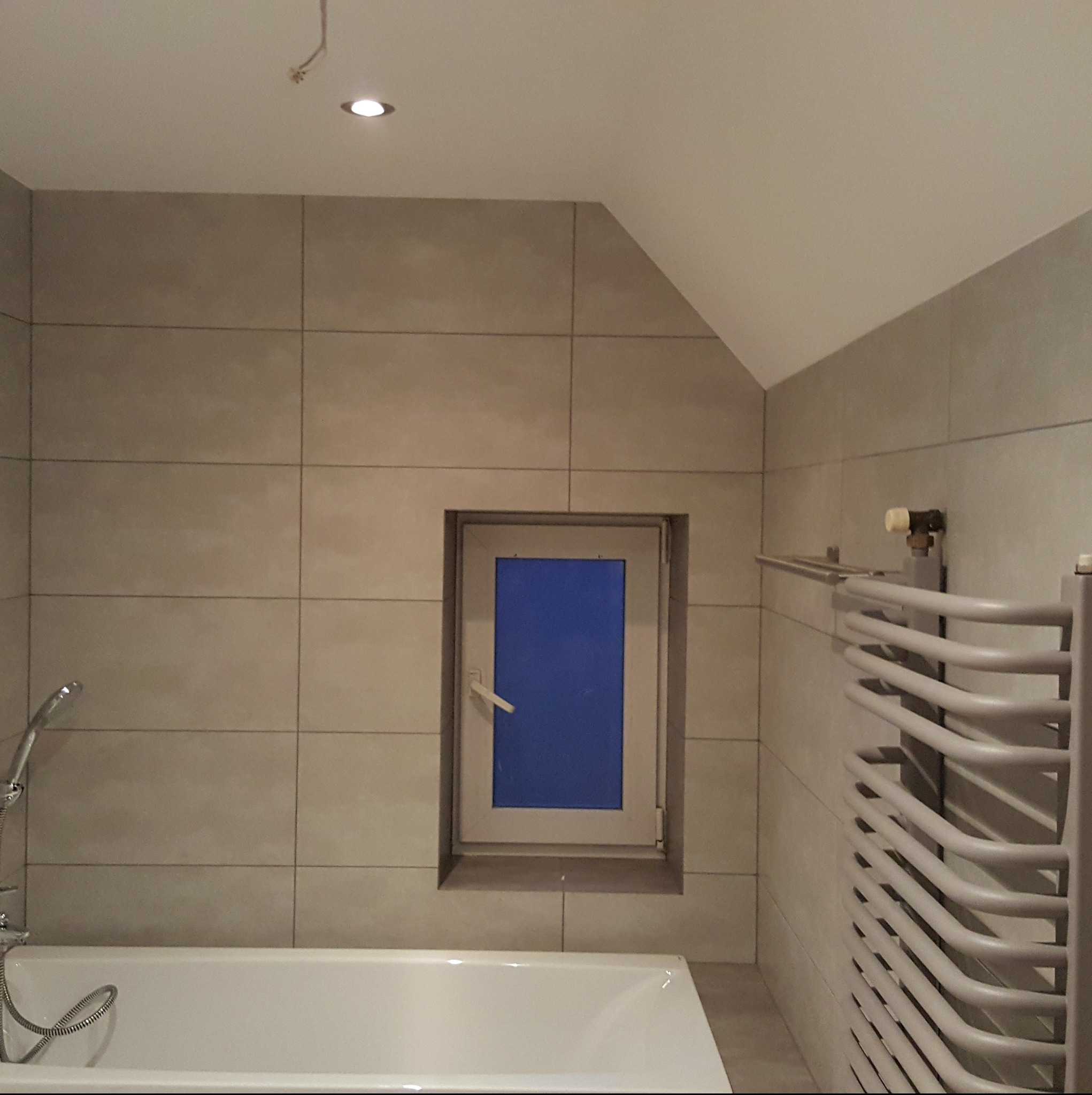 Wykończona łazienka z beżowymi płytkami, białą wanną i grzejnikiem drabinkowym. Okno z niebieską szybą. Minimalistyczny design, neutralna kolorystyka.