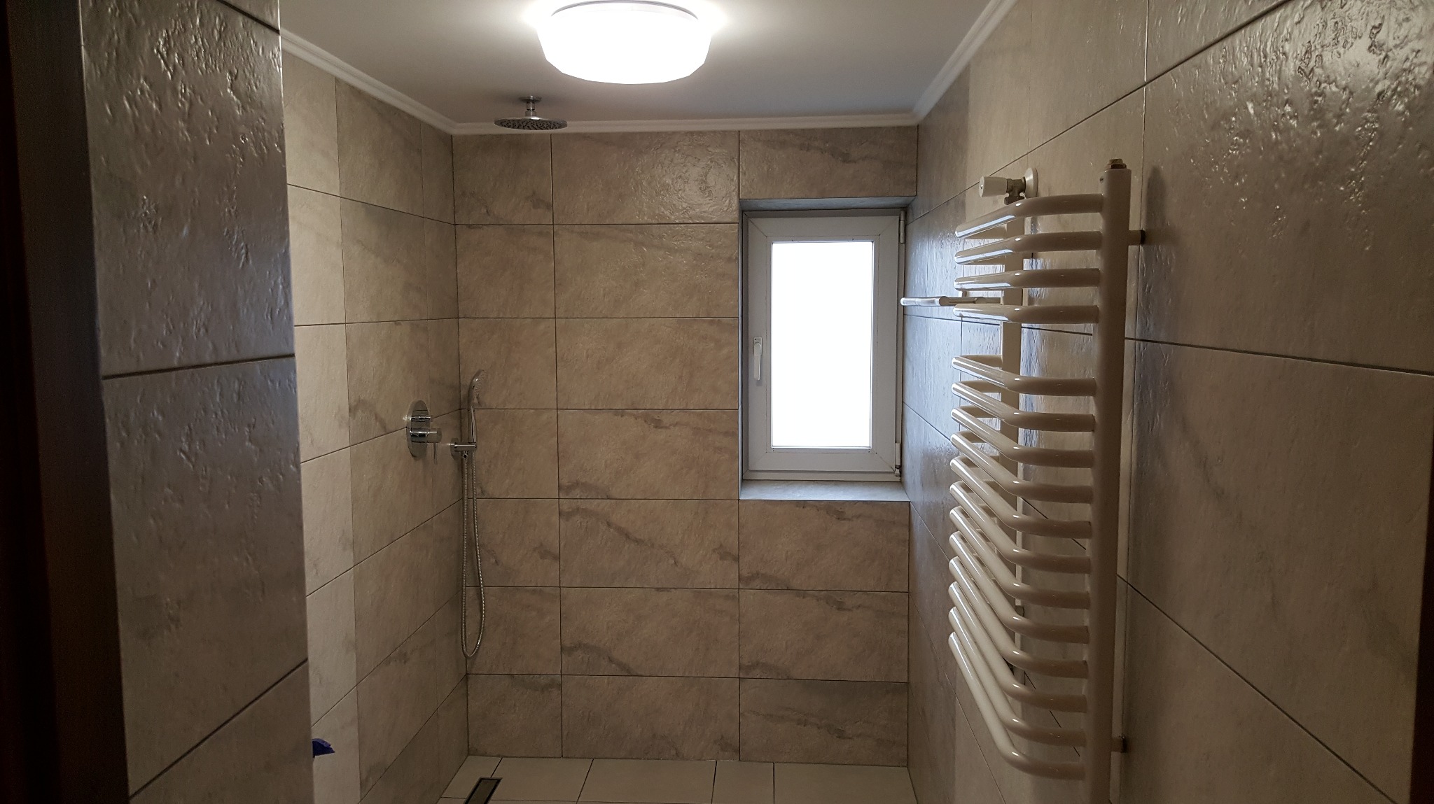 Beżowa łazienka z kabiną prysznicową, grzejnikiem drabinkowym i oknem. Płytki imitujące kamień, oświetlenie sufitowe.