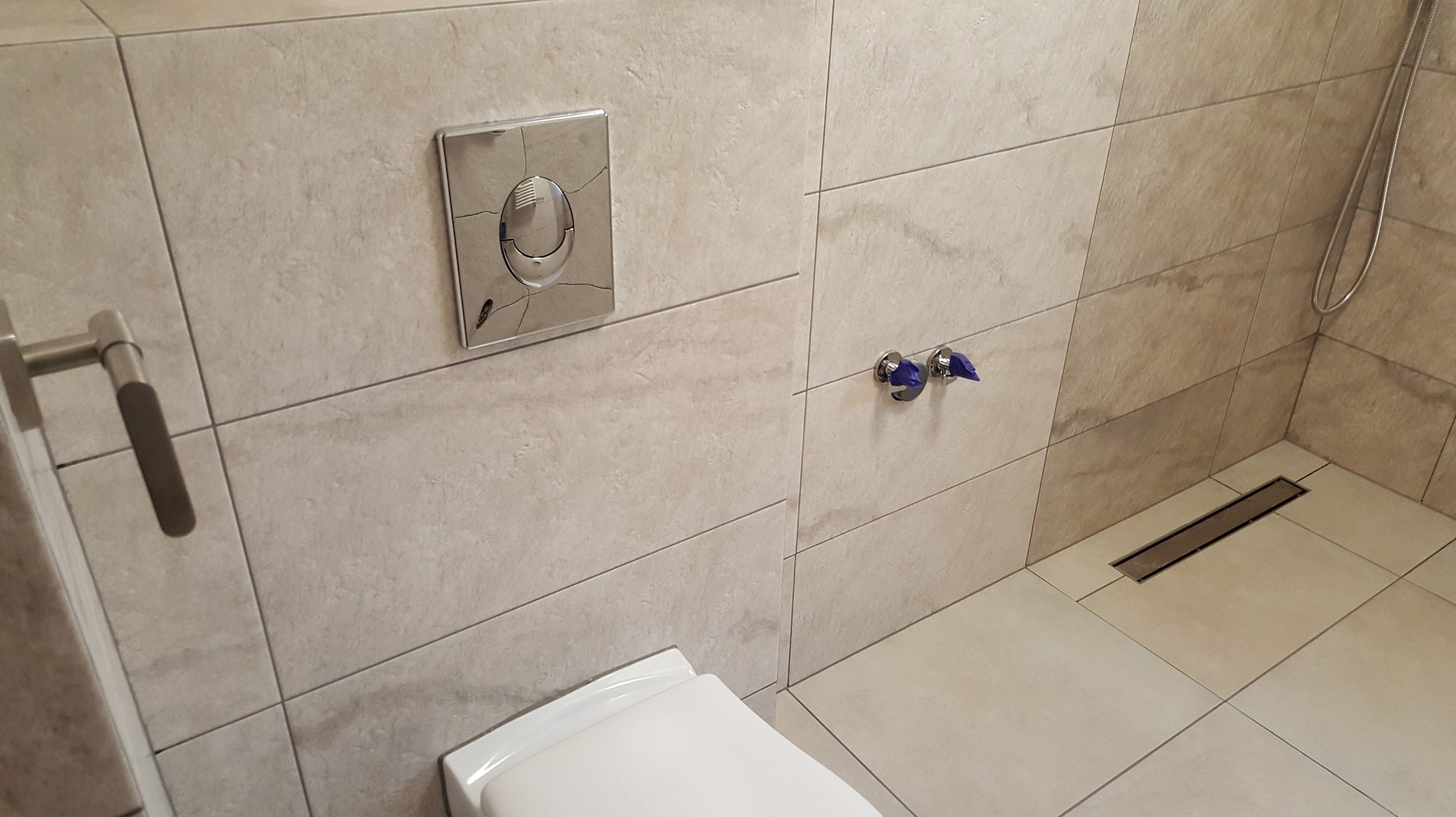 Wykończona łazienka w Lubomierzu: beżowe płytki, podwieszana toaleta z chromowanym przyciskiem, odpływ liniowy i bateria umywalkowa z niebieskim zabezpieczeniem.