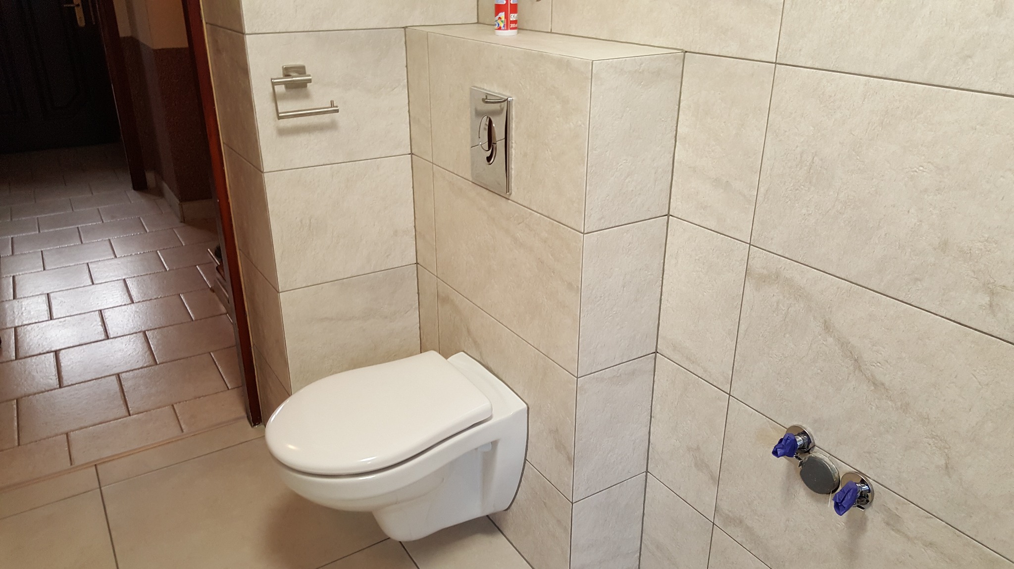 Nowoczesna, podwieszana toaleta w wykończonej łazience z beżowymi płytkami imitującymi kamień. Widoczne chromowane elementy i akcesoria łazienkowe.