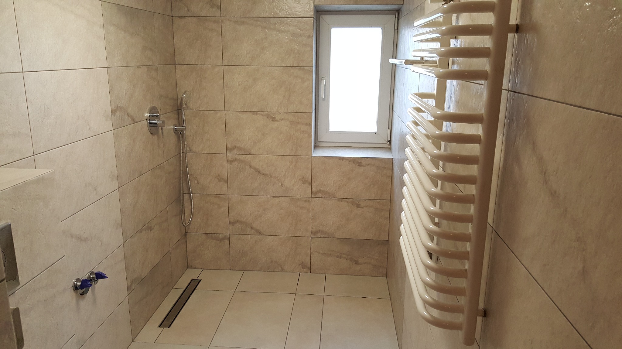 Jasna łazienka po remoncie: beżowe płytki imitujące kamień, grzejnik drabinkowy, chromowana armatura i okno z białymi ramami. Minimalistyczny design.