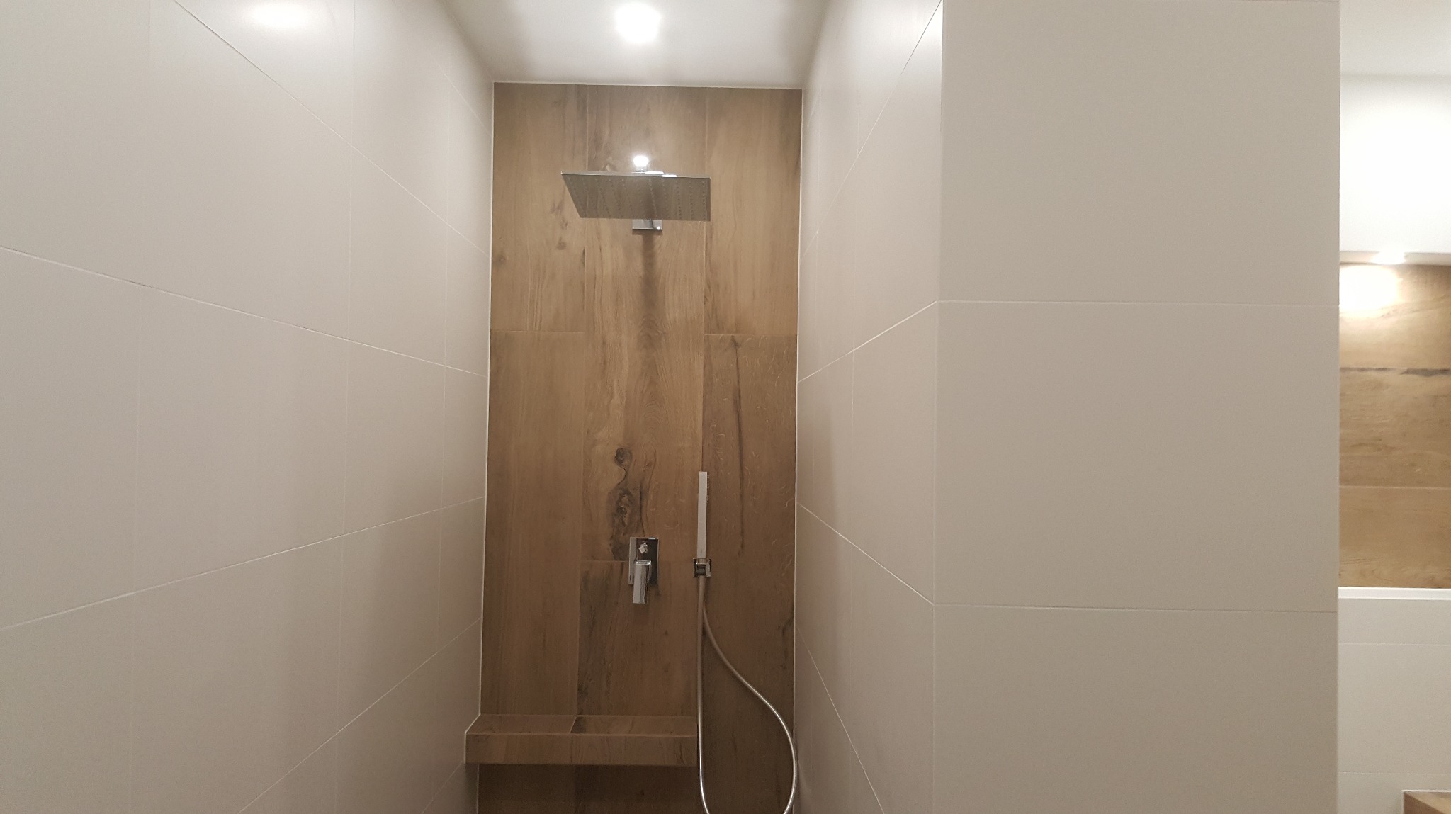 Nowoczesna kabina prysznicowa z deszczownicą i panelami imitującymi drewno, otoczona jasnymi, dużymi płytkami. Minimalistyczny design, stonowana kolorystyka.