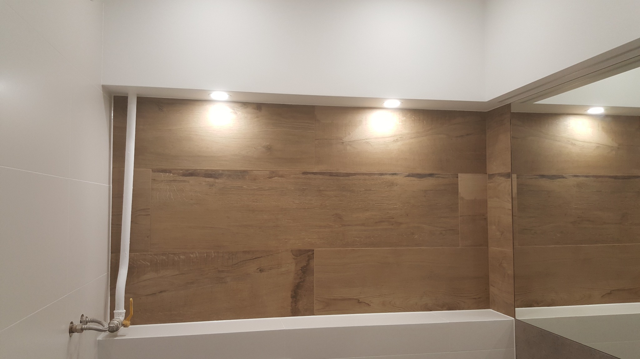 Wykończona łazienka z drewnopodobnymi płytkami, oświetleniem punktowym i białymi elementami. Widoczna instalacja wodna i fragment lustra. Minimalistyczny design.