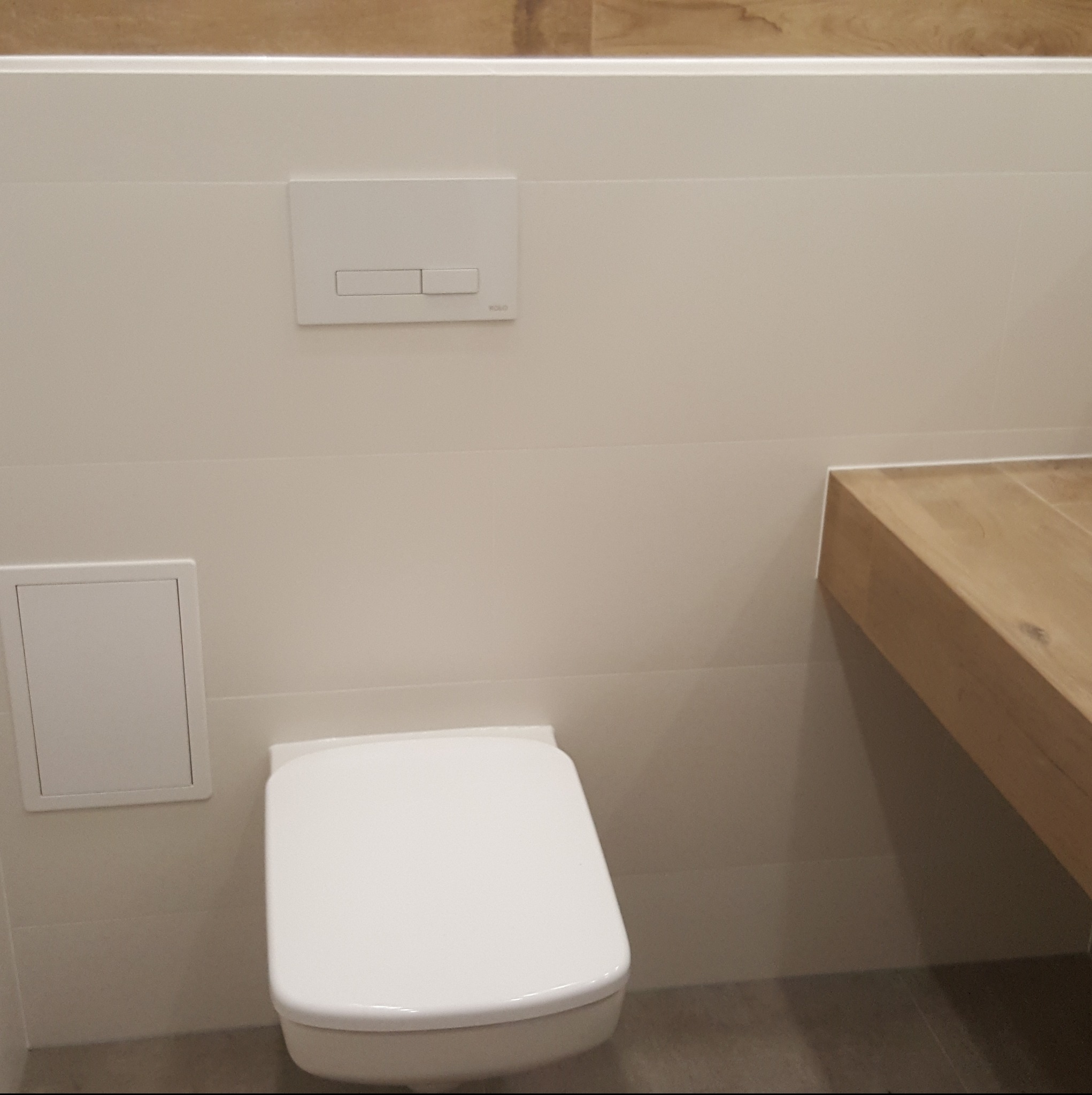 Nowoczesna toaleta podwieszana z białymi płytkami i drewnianym blatem. Minimalistyczny design, czysta linia, widoczny przycisk spłukujący.