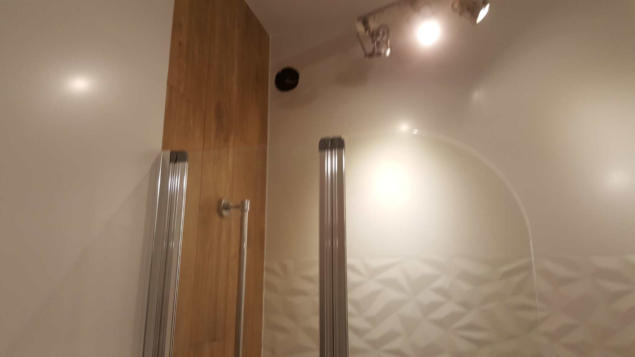 Nowoczesna kabina prysznicowa z drewnianym panelem i geometrycznymi płytkami w łazience. Widoczne oświetlenie punktowe.