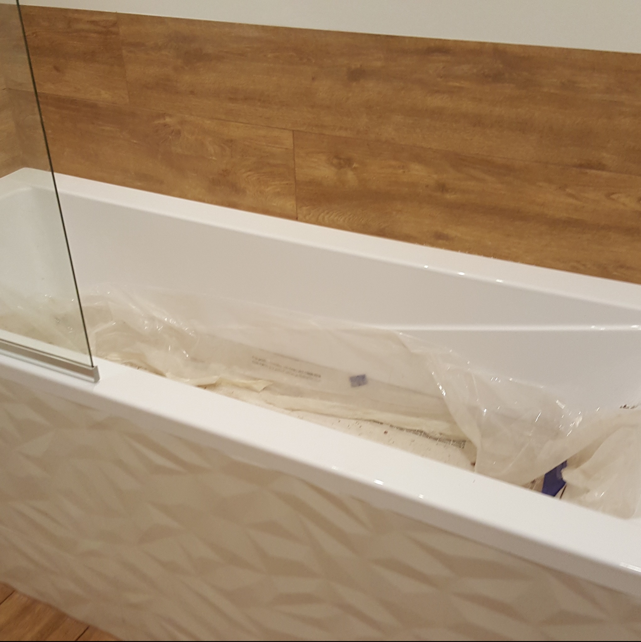 Biała wanna z geometrycznym wzorem na froncie, zabezpieczona folią ochronną. W tle drewnopodobne płytki ścienne i fragment szklanego parawanu.