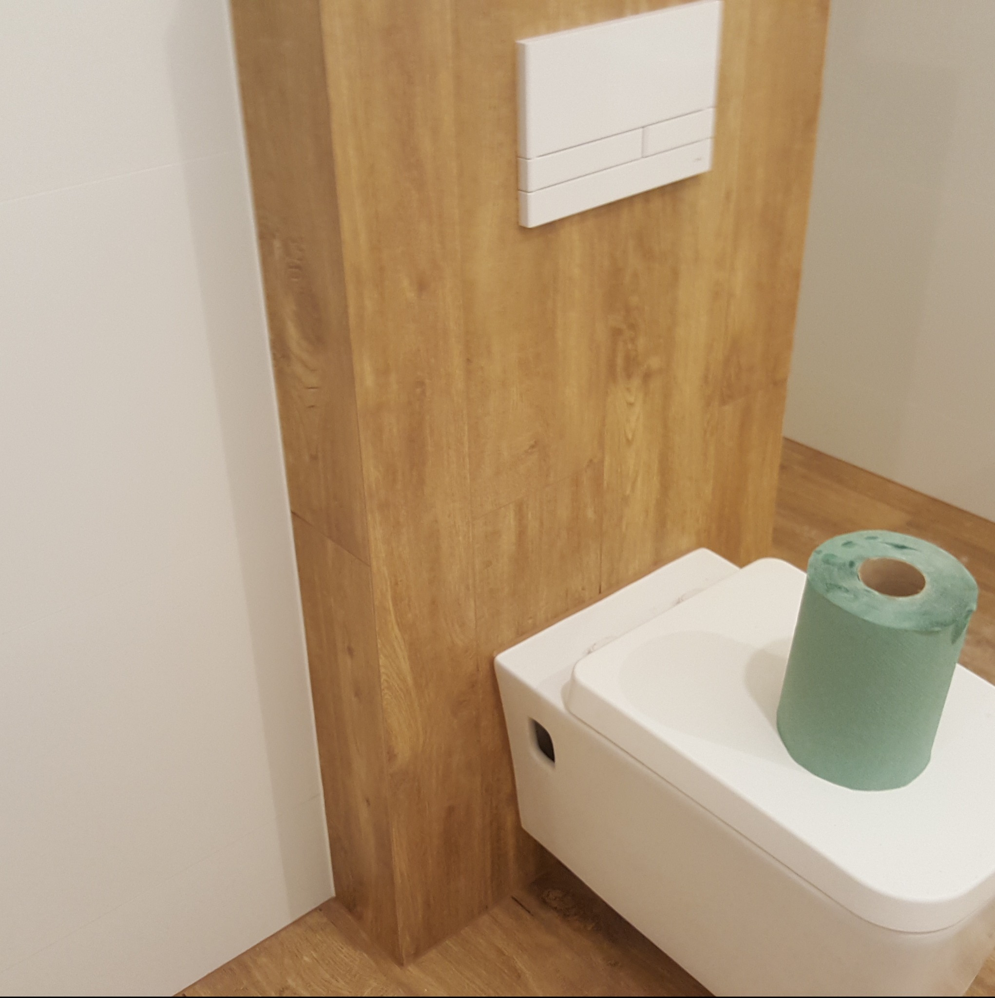 Nowoczesna toaleta podwieszana z drewnopodobną obudową stelaża i zielonym papierem toaletowym. Minimalistyczny design, jasne kolory, wykończenie z dbałością o detale.