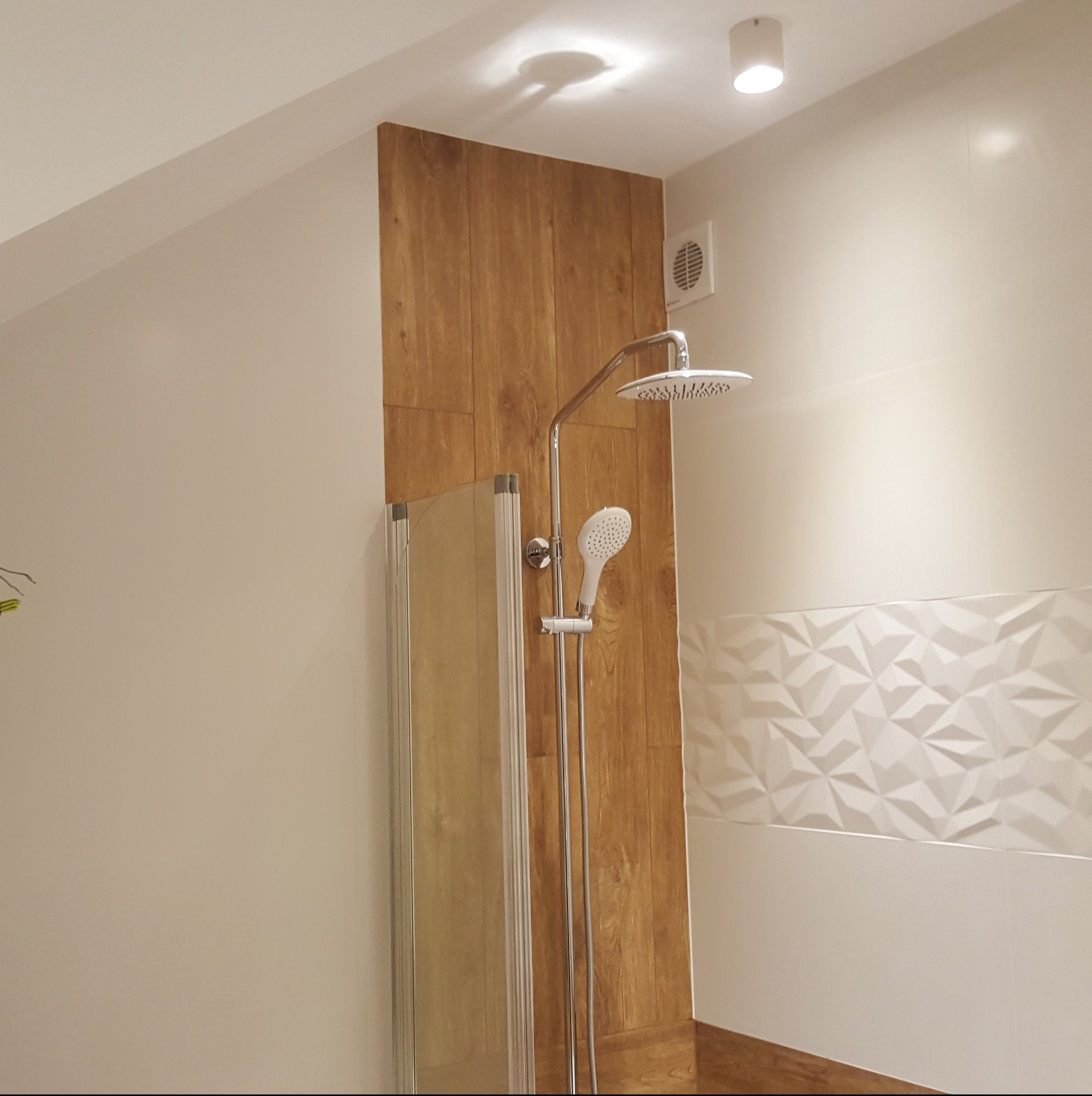 Nowoczesna kabina prysznicowa z deszczownicą i panelami imitującymi drewno. Białe, trójwymiarowe płytki dodają elegancji. Widoczny wentylator i oświetlenie sufitowe.