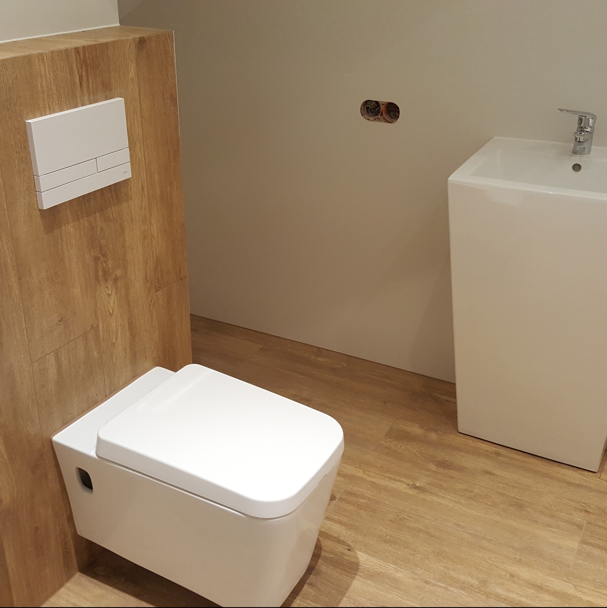 Nowoczesna, podwieszana toaleta z drewnianą obudową i minimalistyczną umywalką w łazience wykończonej panelami podłogowymi imitującymi drewno.