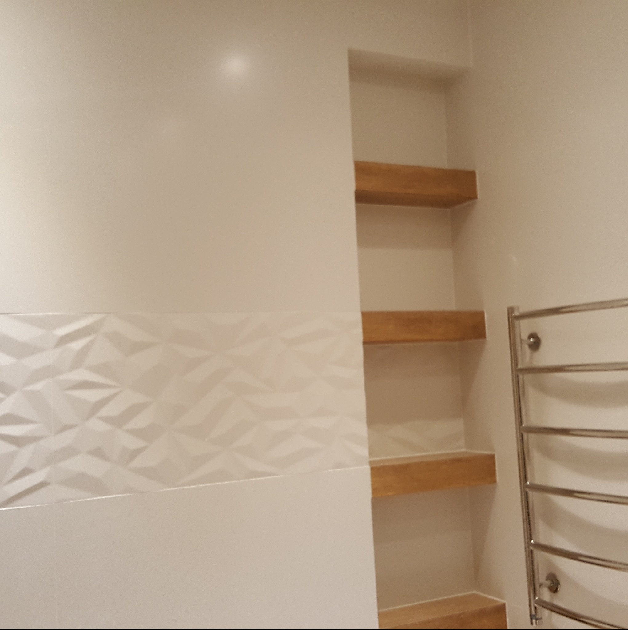 Wykończona łazienka w Lubomierzu: białe płytki 3D, drewniane półki we wnęce i grzejnik drabinkowy. Minimalistyczny design, ciepłe oświetlenie.