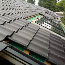 Układanie ciemnoszarej dachówki ceramicznej wokół okien dachowych, widoczne membrany dachowe i drewniane łaty.