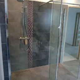 Szara łazienka z kabiną prysznicową typu walk-in, mozaikowy pas dekoracyjny na ścianie i odpływ liniowy w podłodze, widok na drzwi do pokoju.