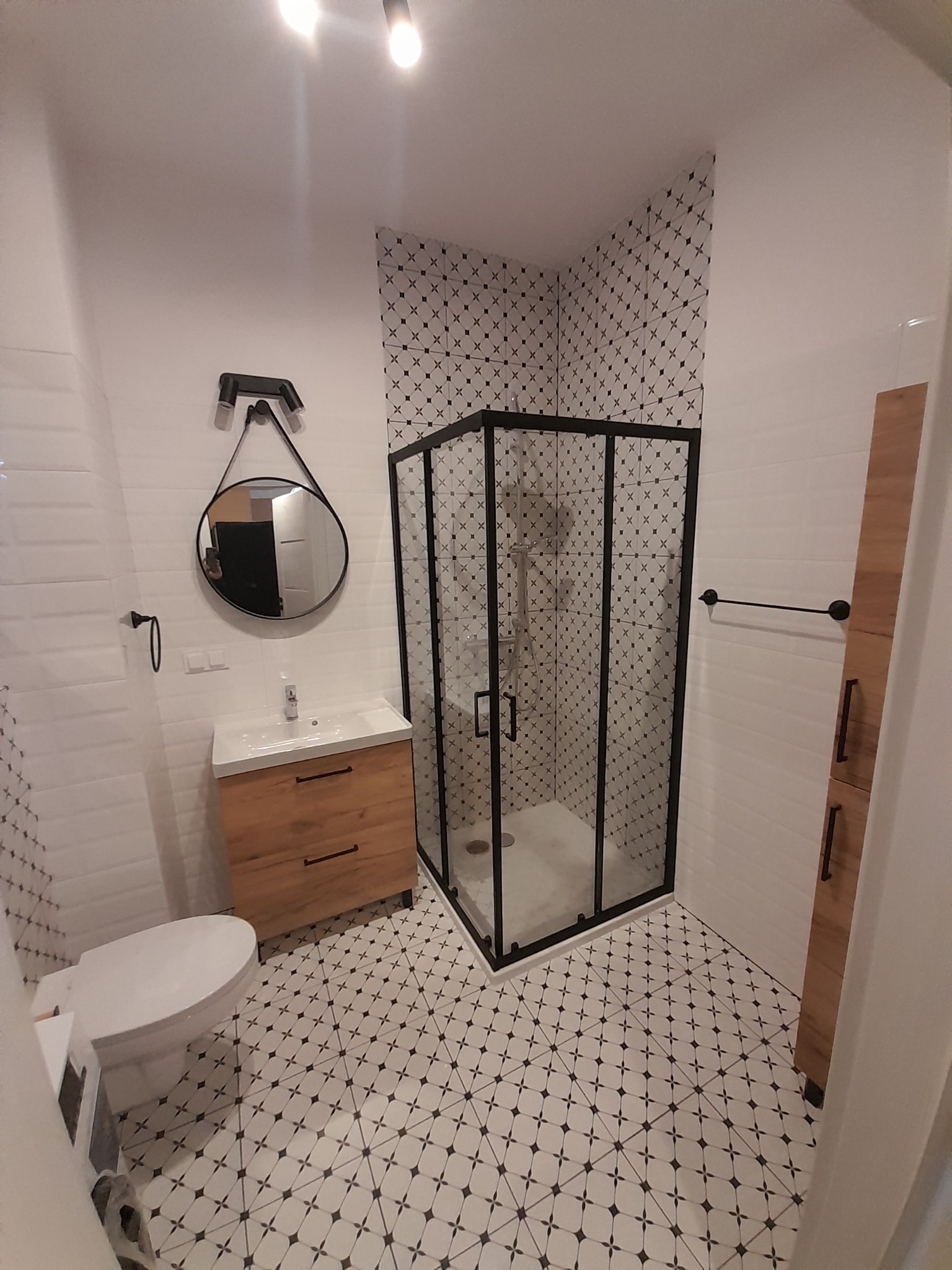 Nowoczesna łazienka z kabiną prysznicową w czarnej ramie, umywalką z drewnianą szafką i okrągłym lustrem na tle białych płytek z czarnym wzorem krzyży.