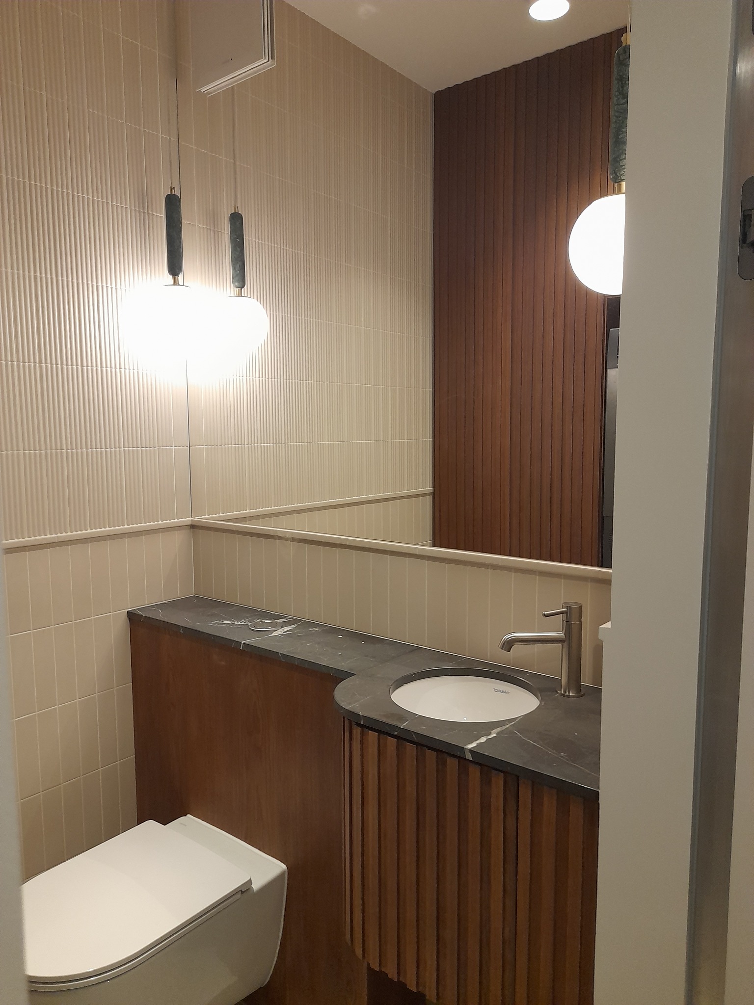 Nowoczesna łazienka z beżowymi płytkami, drewnianą zabudową umywalki z czarnym blatem i wiszącą toaletą. Oświetlenie punktowe i lustro.