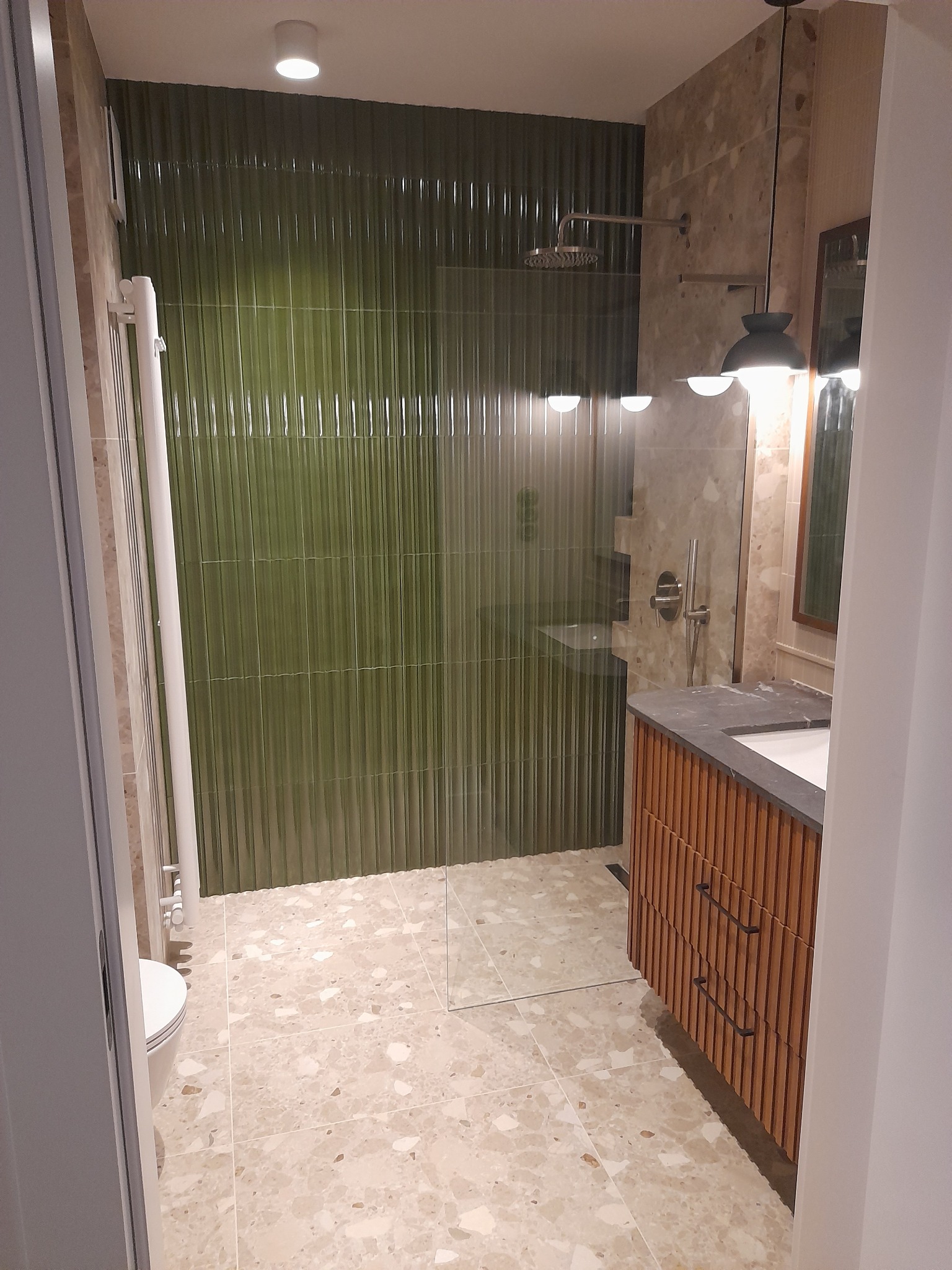 Nowoczesna łazienka z zielonymi płytkami typu jodełka na ścianie prysznicowej, beżową podłogą w terazzo i szafką pod umywalką z lamelami.